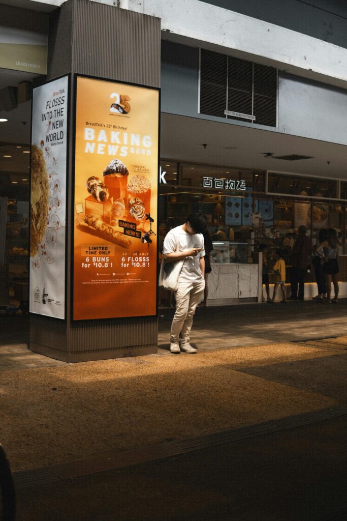 A man stands in sunlight near a bakery advertisement in a bustling urban setting.